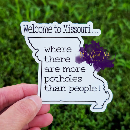A die-cut Missouri-shaped sticker  featuring the phrase “Welcome to Missouri… where there are more potholes than people!” 