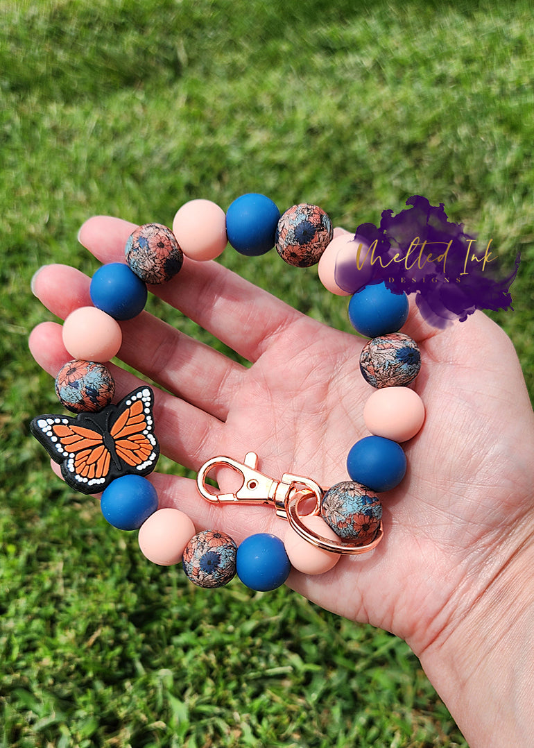 Photo is a wristlet that consists of alternating silicone beads in solid navy blue and light pinkish/orangish color and has blue and burnt orange print silicone bead. Has an orange monarch butterfly and has a rose gold keyring. 