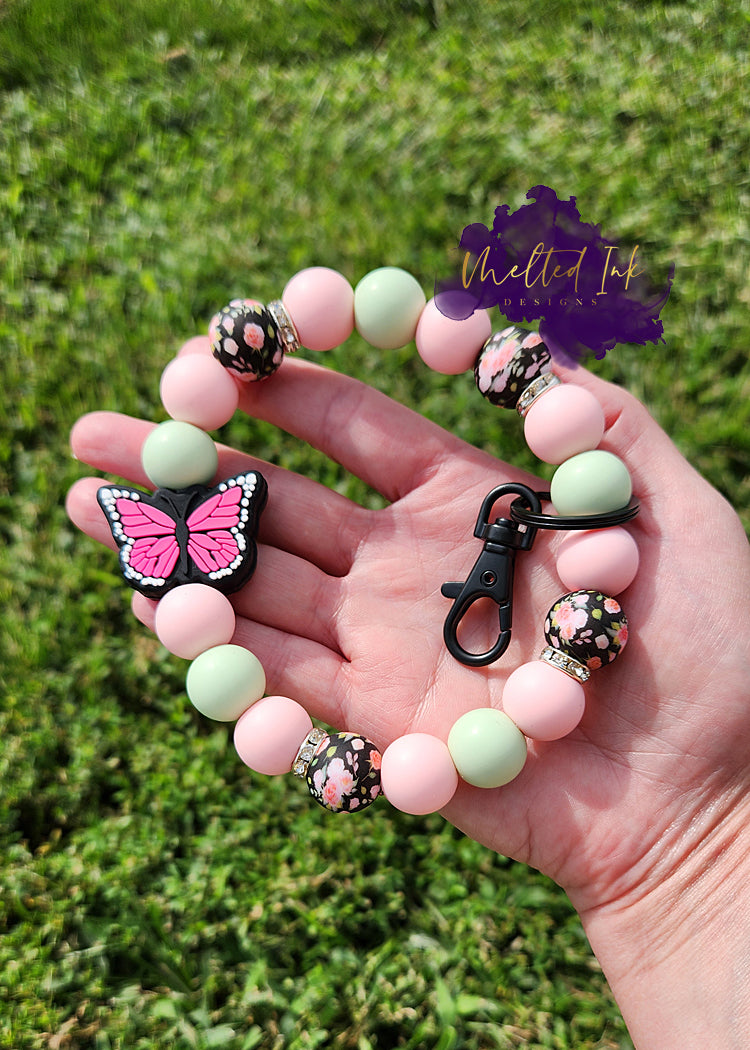 Photo is a keychain wristlet that includes several 15 mm silicone beads that are pink floral print with light pink and mint. Includes a pink monarch butterfly focal bead and has a black keyring. 