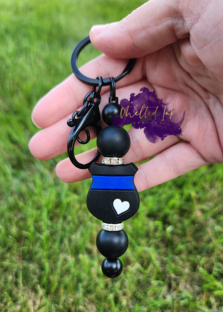 Photo consists of black beaded keychain bar that has a thin blue line badge with a white heart on it. It is accented with black silicone beads and rhinestone spacers on top and bottom of the badge. 