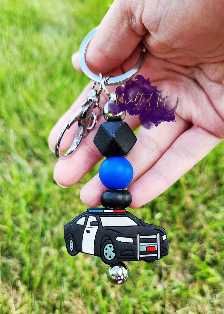 photo is a silver beadable keychain bar that consists of alternating black and blue solid silicone beads above a police cruiser focal bead. 