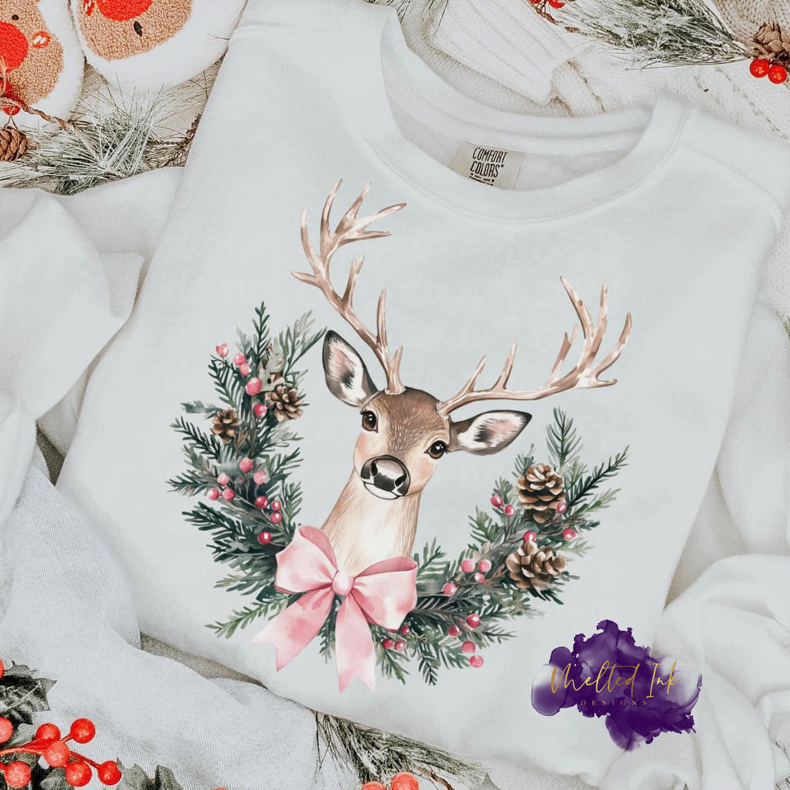 Photo is a mock up of design and consists of a deer behind half of a Christmas wreath that has a pink bow on it. 