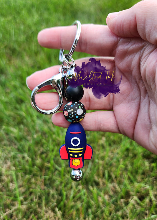 photo is a silver beadable keychain bar that has a solid black and planet silicone beads. also has a spaceship on the bottom