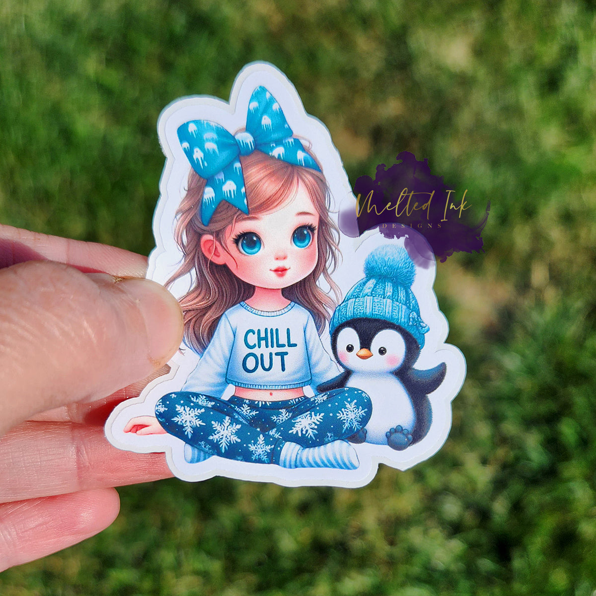 Photo is of a sticker that has a little girl with a bow in her hair, snowflake pants and a shirt that says chill out. Next to her is a little penguin in a pom pom hat. 