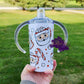 Photo consists of a kids sippy cup that converts into a tumbler. The design on the cup consists of  santa face mugs filled with marshmellows. Background has marshmellows, candy canes, and little dots and stars. 