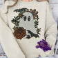 Image of a friendly ghost made entirely of sequins, surrounded by bronze and black floral details, green leaves, and two small bats. The design sparkles under the light, blending spooky and elegant elements in an autumn-themed style.