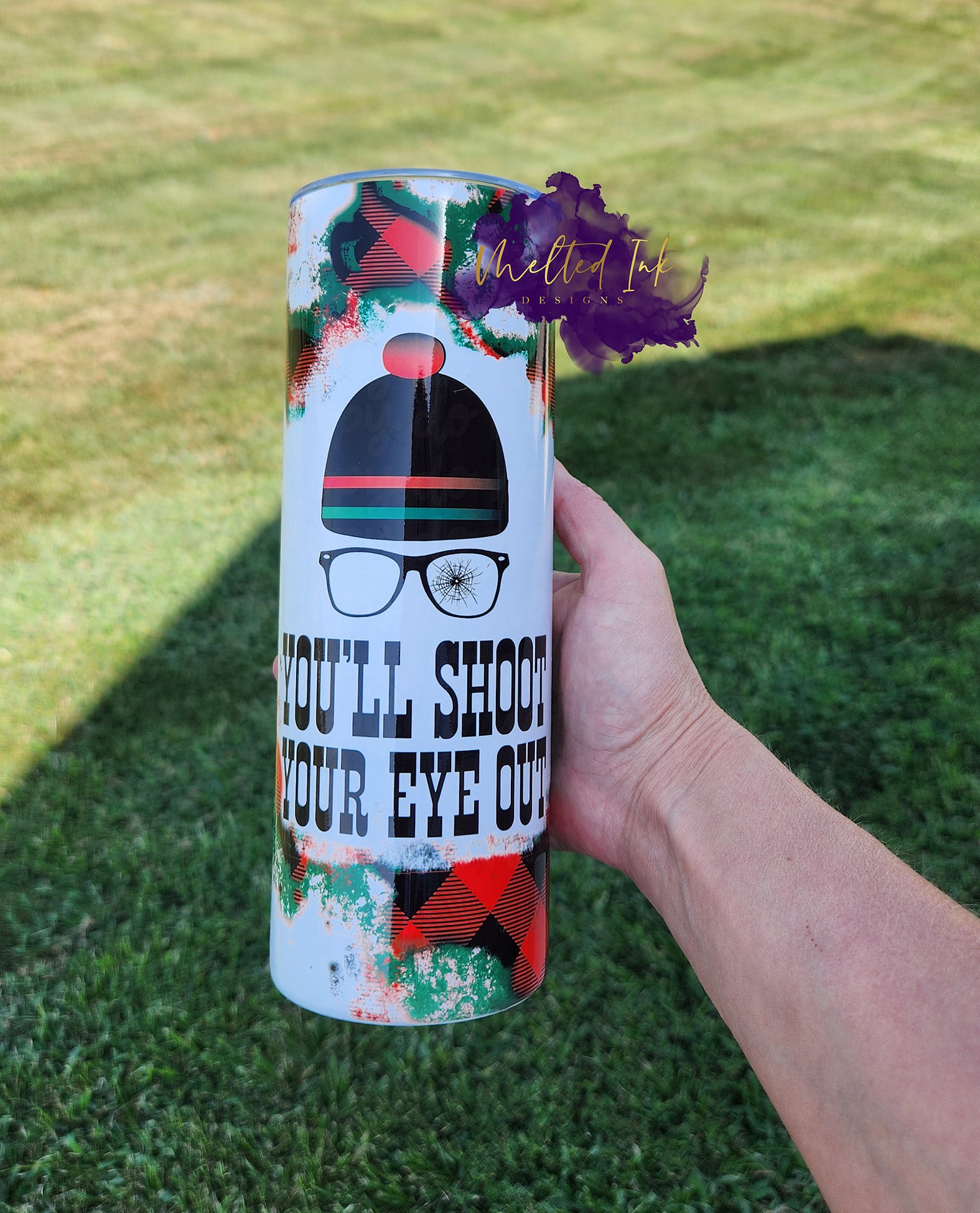 Photo is a 20oz tumbler that has a faded red and black checkered background and has a picture of a hat with eye glasses with the saying, I'll shoot your eye out. 