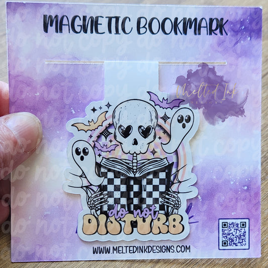 his magnetic bookmark features the phrase "Do Not Disturb," paired with a whimsical illustration of a skeleton engrossed in a book. Little ghosts and bats float around the skeleton, adding a fun, spooky touch to the design.