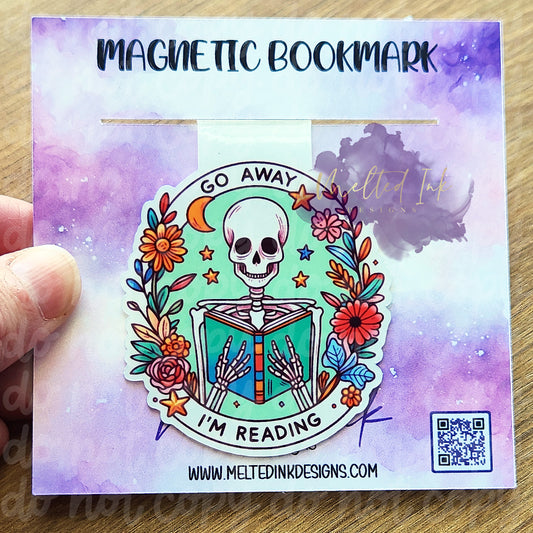 This magnetic bookmark features the phrase "Go Away, I'm Reading," complemented by a playful illustration of a skeleton deeply engrossed in a book. Surrounding the skeleton are vibrant flowers, adding a cheerful touch to the design.