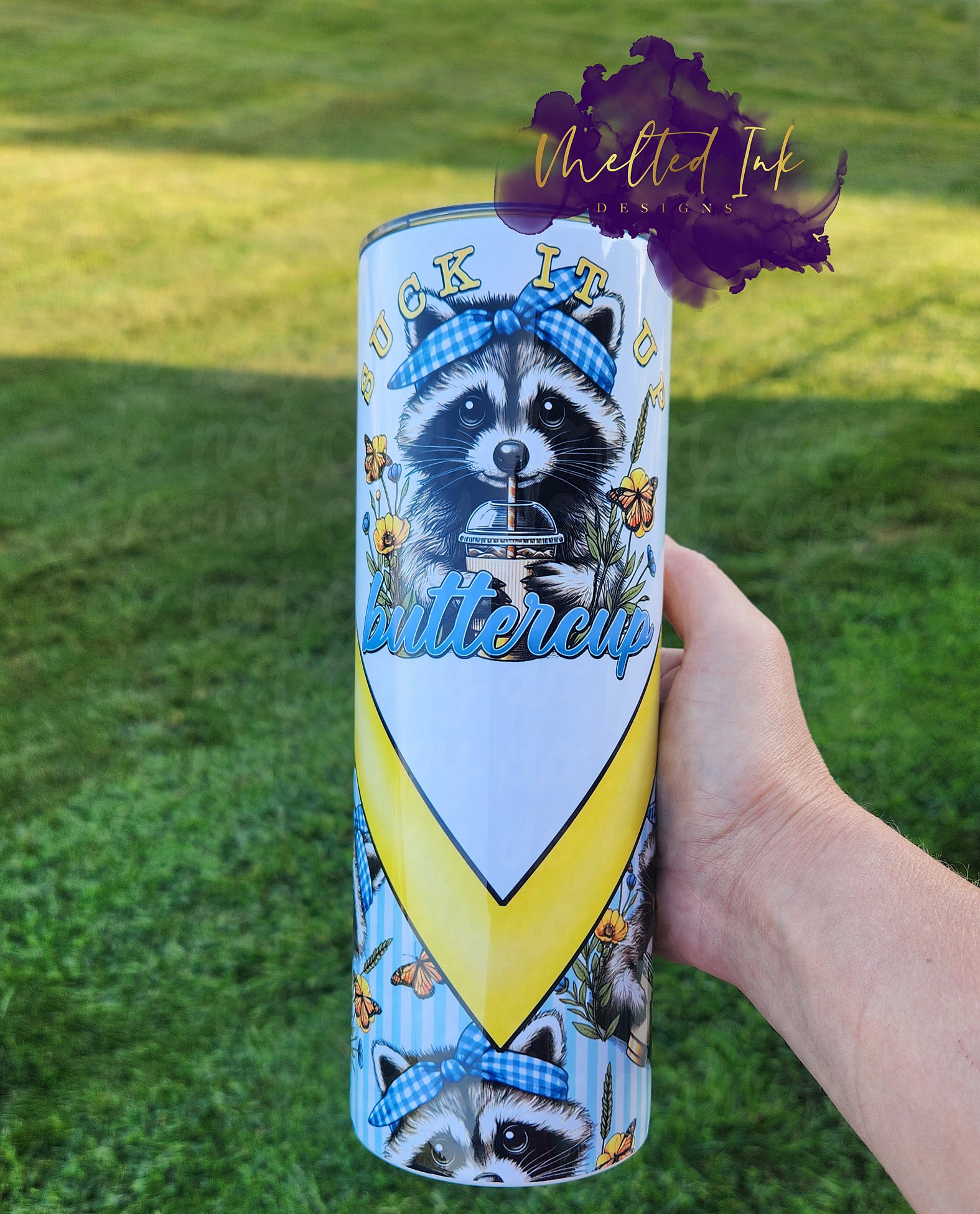 The photo features a charming 20oz tumbler showcasing an adorable baby raccoon sipping coffee through a straw. The raccoon sports a cute blue and white checkered headband, adding a playful touch to its look. The cup is emblazoned with the phrase "Suck it up, Buttercup," which adds a fun and motivational twist.