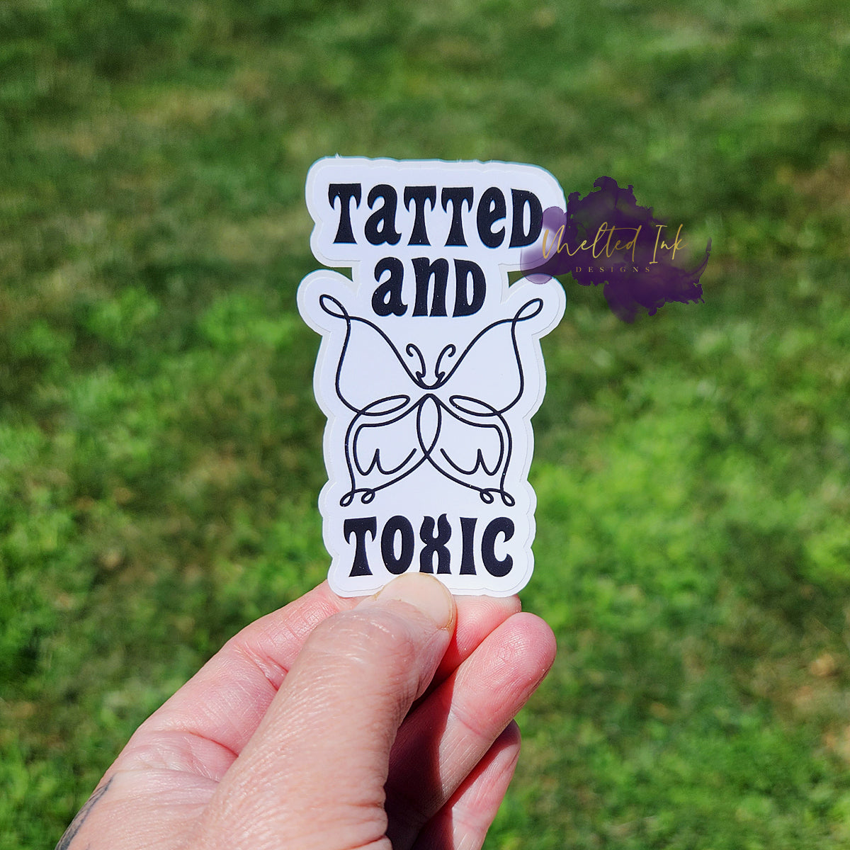 Photo is of a sticker that says tatted and toxic with an outline of a butterfly. Ink is black. 