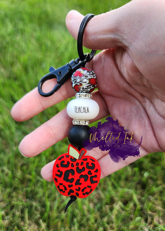 Photo consists of a teacher keychain and has the following beads: red apple with black leopard print, solid black, and apple leopard print silicone bead as well as a silicone spacer bead that says teacher. The beads are accented with rhinestone spacers. 