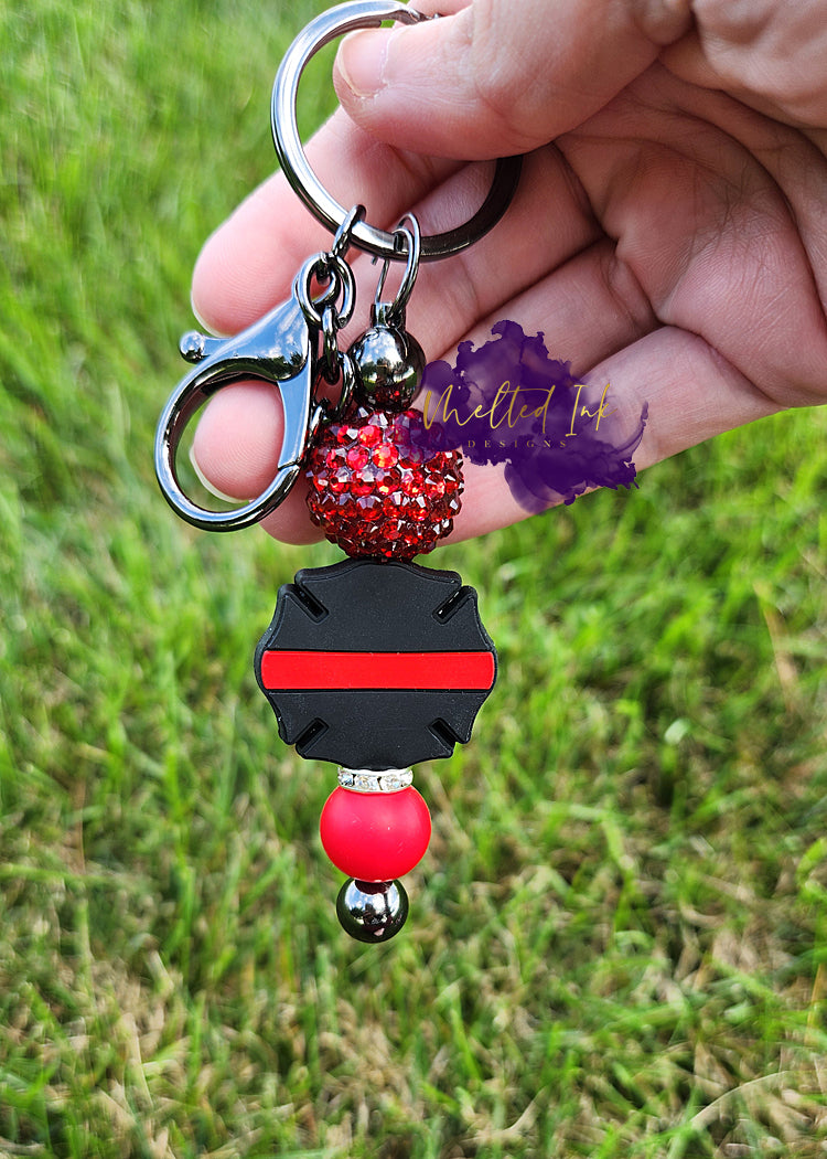 Photo consists of a dark gray beadable keychain bar that has a thin red line firefighter badge. It is accented with an acrylic spacer, a solid red silicone bead and rhinestone spacer.