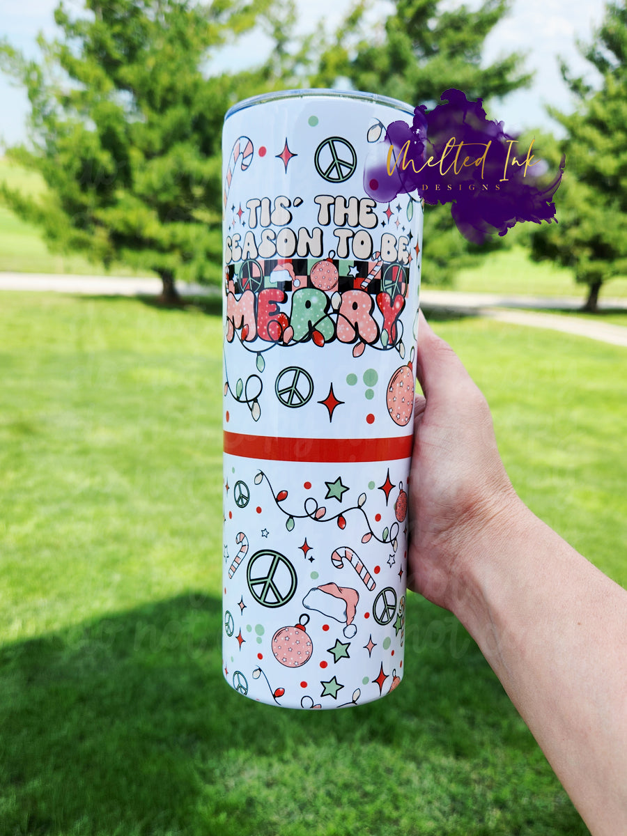 The photo features a festive 20oz Christmas tumbler adorned with the cheerful phrase "Tis the season to be merry." The design incorporates vibrant red, light pink, and light green colors, creating a joyful holiday spirit. It includes playful elements like candy canes, a peace sign, Santa hats, and colorful Christmas lights. Scattered throughout are stars and polka dots in light green and red, adding to the overall whimsy. 