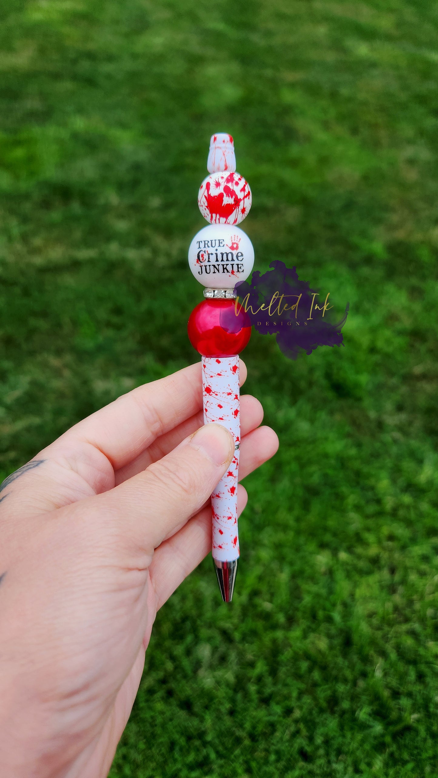 Photo is of a printed blood splatter beaded ink pen. It has an acrylic bead that says True Crime Junkie. On top of of that bead is a bloody hand print wooden bead. On the bottom is a red acrylic bead. Ink is black. 