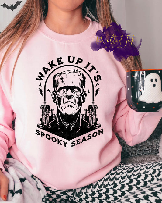 Photo is a mock up of the design. Shirt design color is black and pressed on a pink sweatshirt. Design says: Wake Up It's Spooky Season with picture of Frankenstein. 
