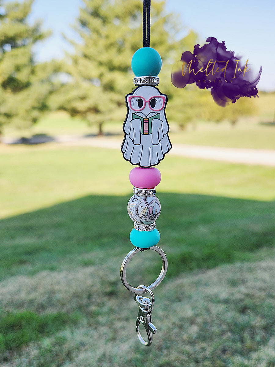 his lanyard features an adorable focal bead of a ghost reading a book, capturing a whimsical and playful vibe. It is accented with light blue and pink silicone beads, along with sparkling rhinestones, adding a fun touch to the design. The lanyard also includes a keyring .