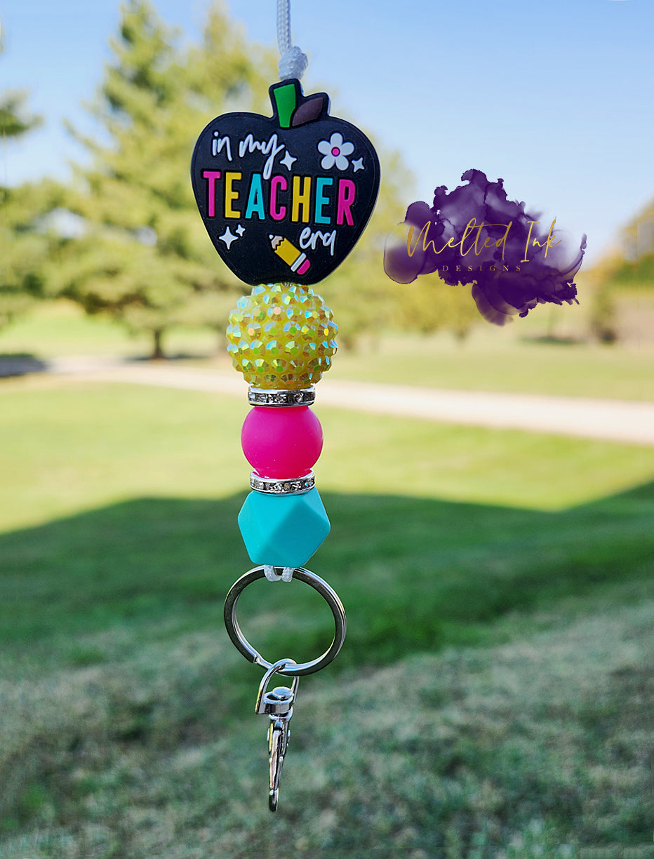 This teacher lanyard features a charming black apple focal bead that says "In My Teacher Era," with the word "Teacher" highlighted in alternating colors of pink, yellow, and light blue. The lanyard includes a cheerful yellow acrylic bead, a pink 15mm silicone bead, and a light hexagon bead, all adding to its vibrant design. It's accented with rhinestones for a touch of sparkle and comes with a keyring 