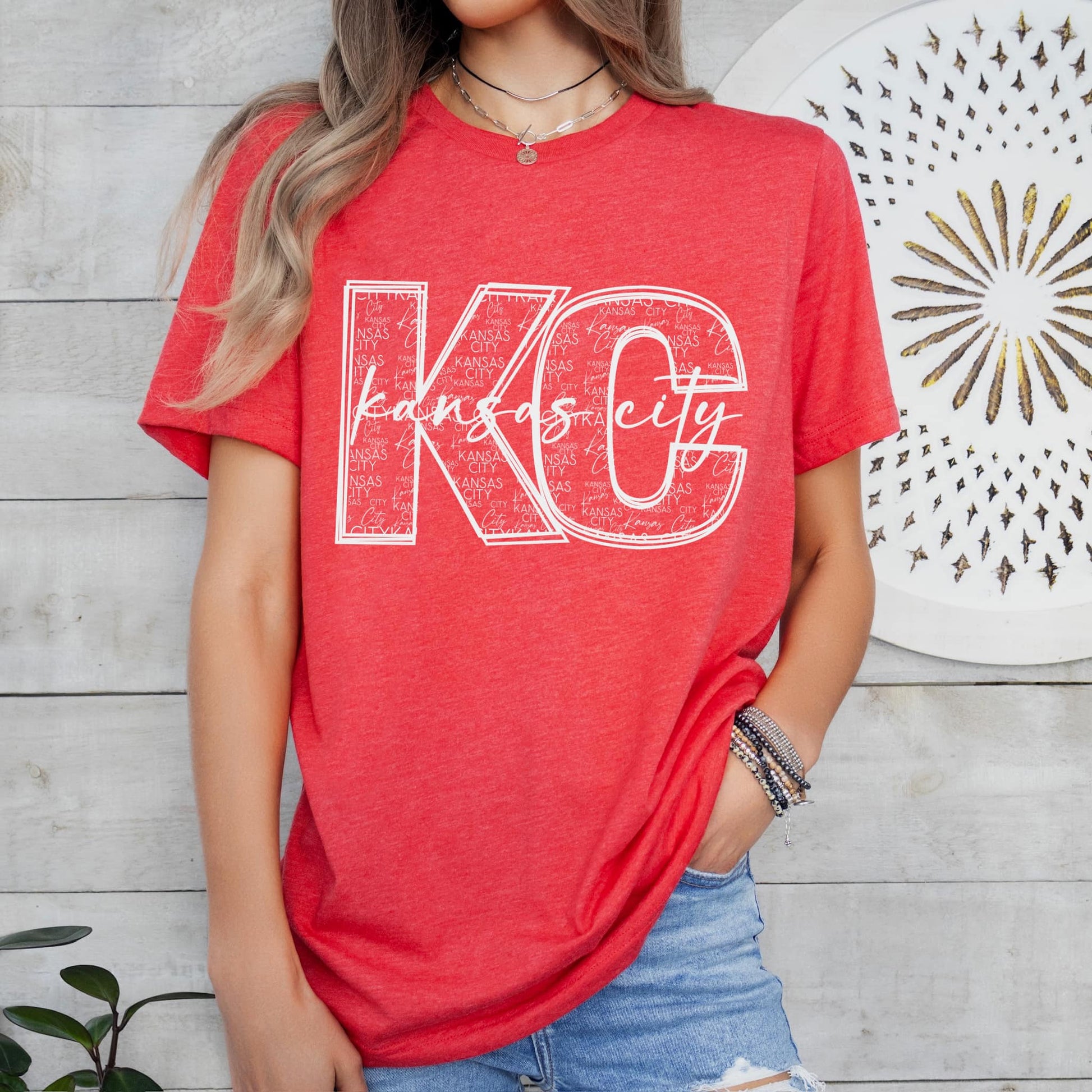 Photo is a mock up of the design. Design color is white and the letters KC with Kansas City written over top of them. Inside the KC letters the words Kansas City is written out several times. Design is pressed on a red shirt. 