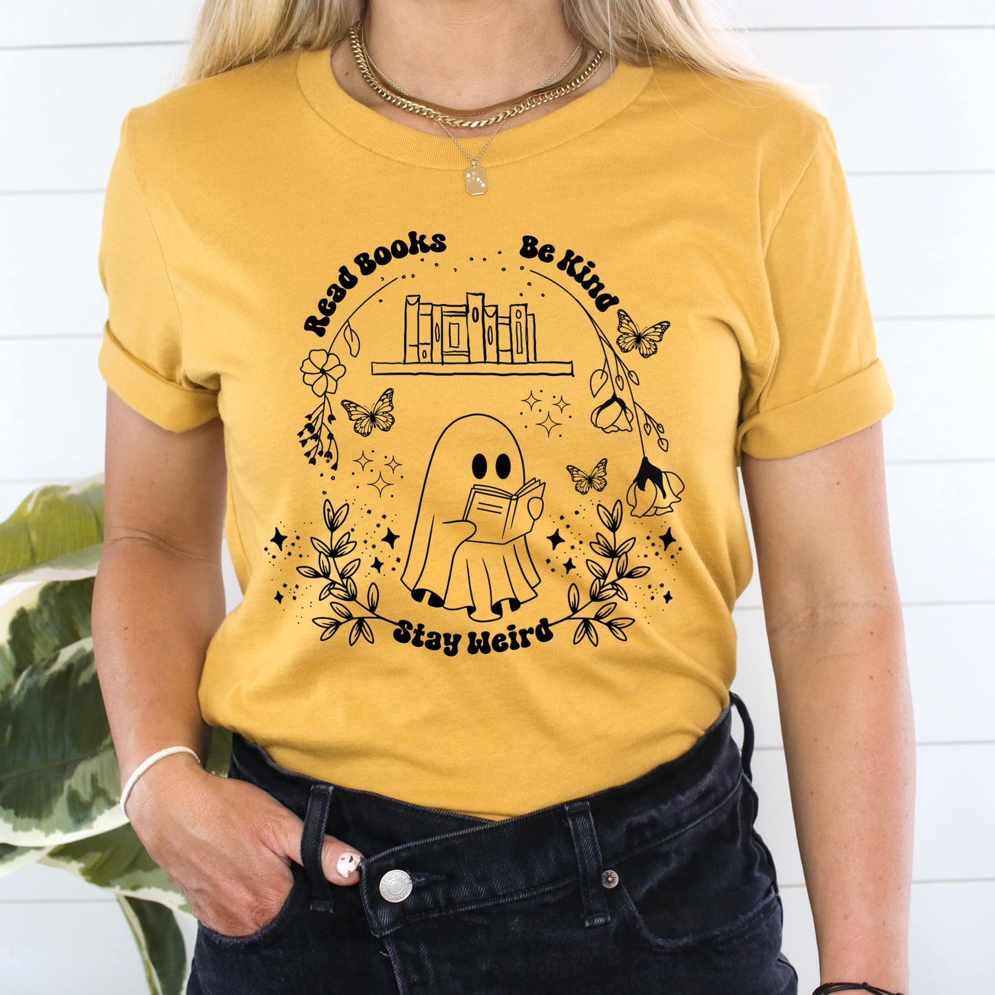 Photo is a mock up of the design. Design print is black and is pressed on a yellow shirt. Design says Read Books Be Kind Stay Weird. Picture is of a ghost reading a book with a bookshelf above it that has books on it. Includes butterflies flowers and leaves. 