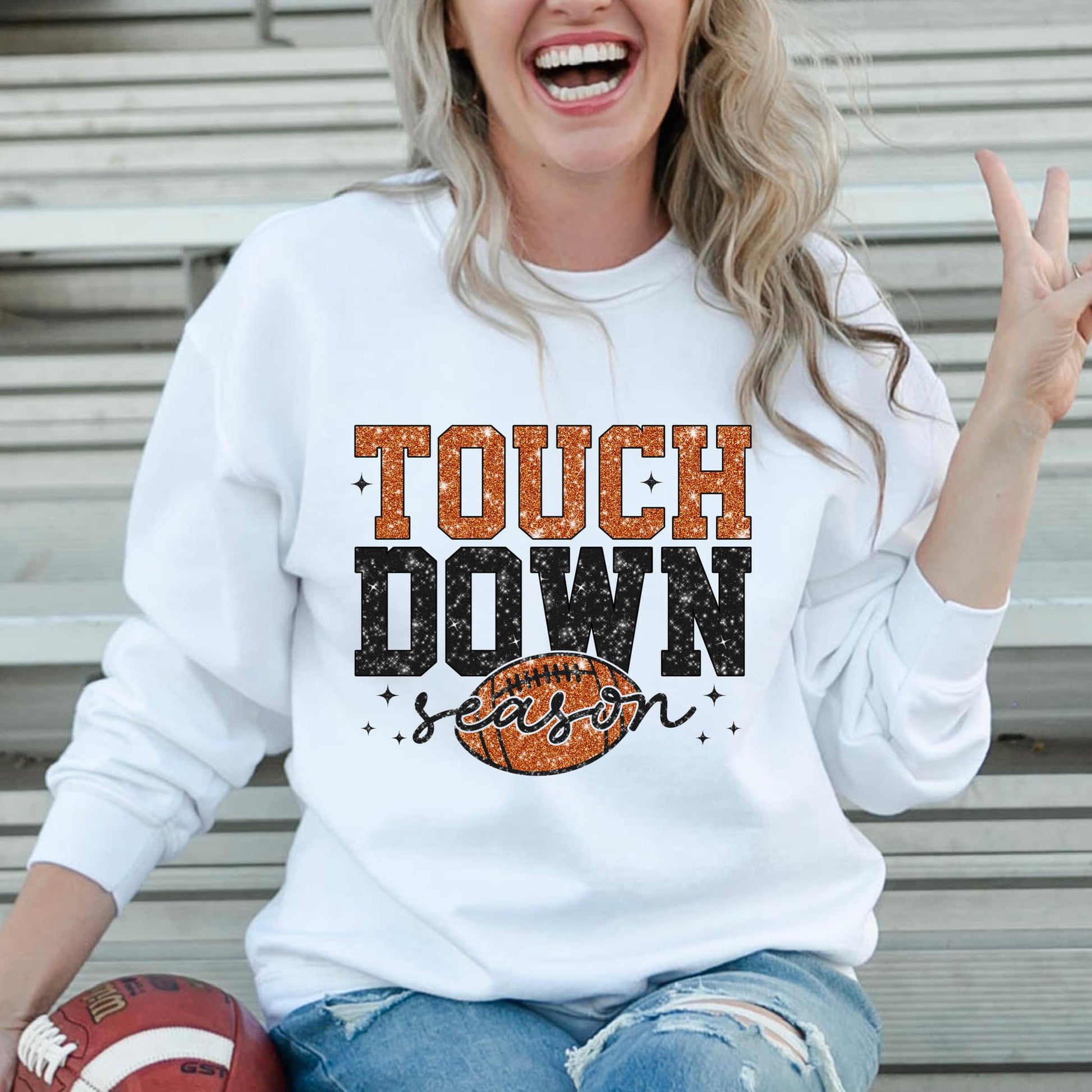 The product photo provided is only a mock up of the design. This design is a faux glitter design in black and brown. It says Touch in brown and Down in Black. These words are stacked on top of each other. The design includes a brown football and the word season is layered on top of the football. Design is on a white sweatshirt.