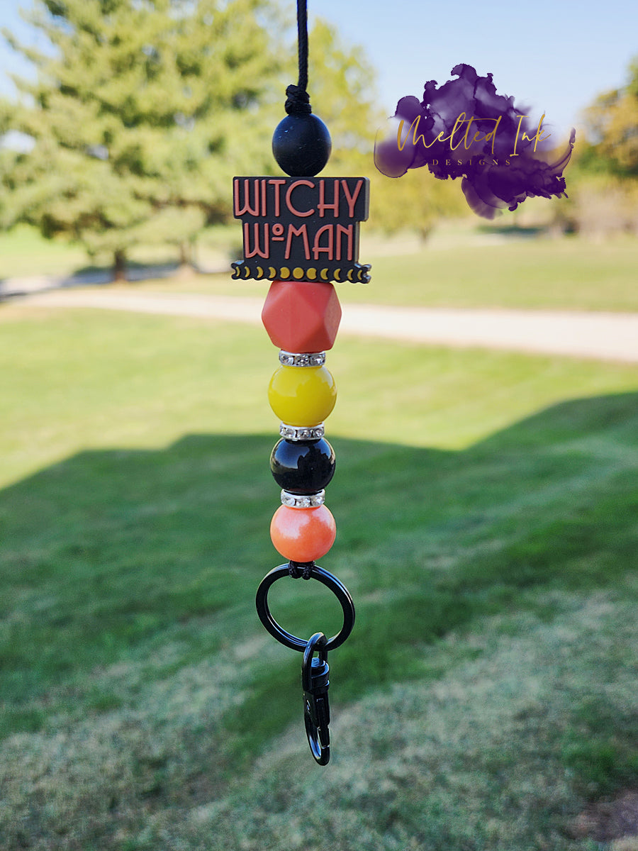 This Halloween lanyard features a focal bead that says "Witchy Woman" in striking black and orange. It includes 15mm silicone beads in vibrant yellow, black, and orange, along with a unique orange hexagon silicone bead. The design is accented with rhinestone spacers for a touch of sparkle and comes with a keyring for convenience. 